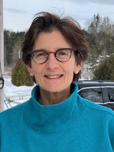 woman in blue shirt, with short brown hair blowing in the wind, wearing glasses, standing outside on a snowy day