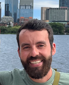 Man with dark hair and beard along Charles River in Cambridge, with Boston in the background