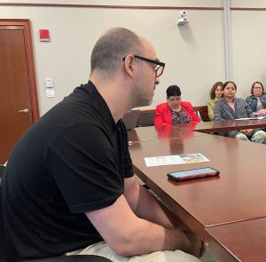 White man in black tshirt and glasses speaks to a group of people assembled at a large table