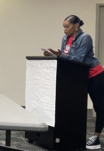 Black woman in jeans jacket and red shirt presenting from a podium at a tenant council meeting