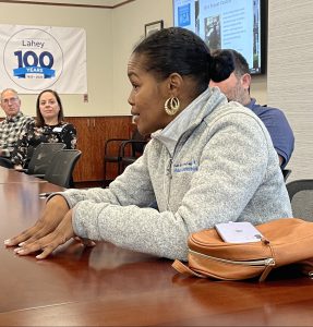 black woman with hair pulled back, wearing gold earnings, speaking at a conference table to other people