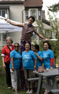 A stiltwalker in a polka dotted vest stands tall behind 5 smiling women, 4 of them are in blue Walk for Affordable Housing shirts.