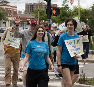 Two young women in blue t-shirts. One holding a Walk for Affordable Housing sign. Behind them, a man in a red baseball hat is waving at the camera and others are walking with signs.