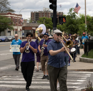 Men in foreground playing horned instruments crossing the street, followed by other people holding signs