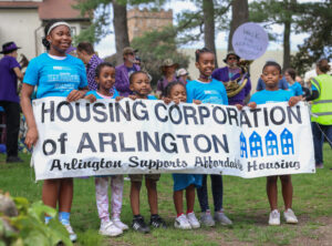 five mostly smiling black children of varying ages and heights are proudly holding a sign that says Housing Corporation of Arlington: Arlington Supports Affordable Housing. They are standing in a field. They are wearing turquoise Walk for Affordable Housing shirts