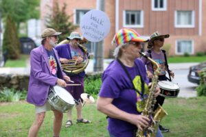 four people wearing purple. Two are playing horn instruments and two are playing drums. They are standing on a lawn. One has a sign that says Walk for Affordable Housing attached to his instrument.