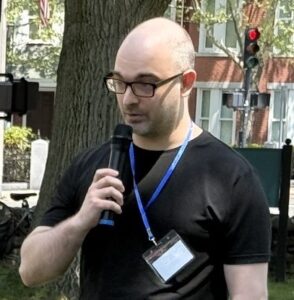 White man in glasses and a black t-shirt standing outside speaking into a microphone