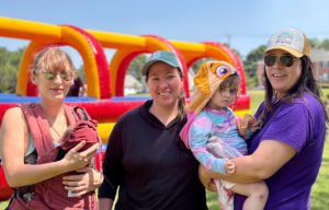 Three white women standing outside in a field on a sunny day. One is holding a baby. Another is holding a toddler.