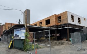 View of building under construction on cloudy day. It has one floor of steel frame and one floor of wooden framing. An elevator shaft sticks up in the middle.